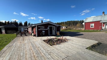 Una terraza de madera que lleva a un edificio con la leyenda 'HOTEL CAFE RESTAURANG', con un cielo despejado y un bosque al fondo.