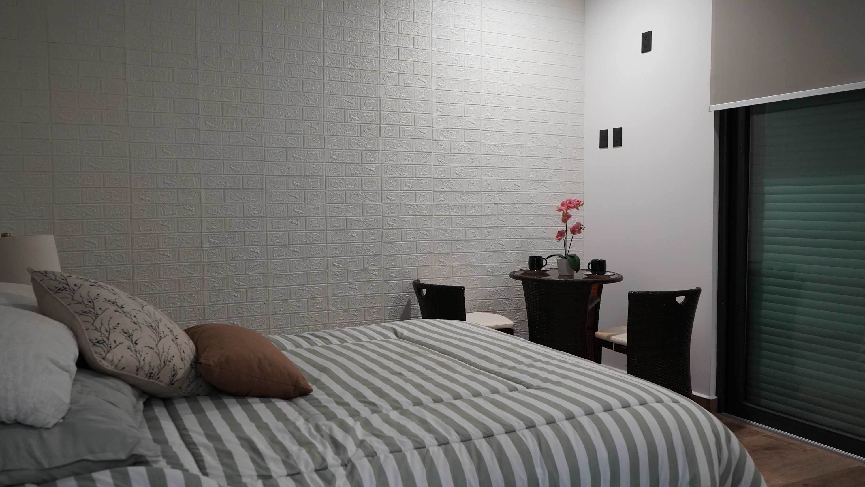 A bedroom with a bed, pillows, a nightstand, a small table with a vase of flowers, and a sliding door.
