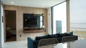 A modern living room with a flat-screen TV mounted on a wooden wall, a sofa with cushions, and a dining area with a table and chairs. - Torre W Beachfront Condo: Ocean View & Pools (Mazatlán)