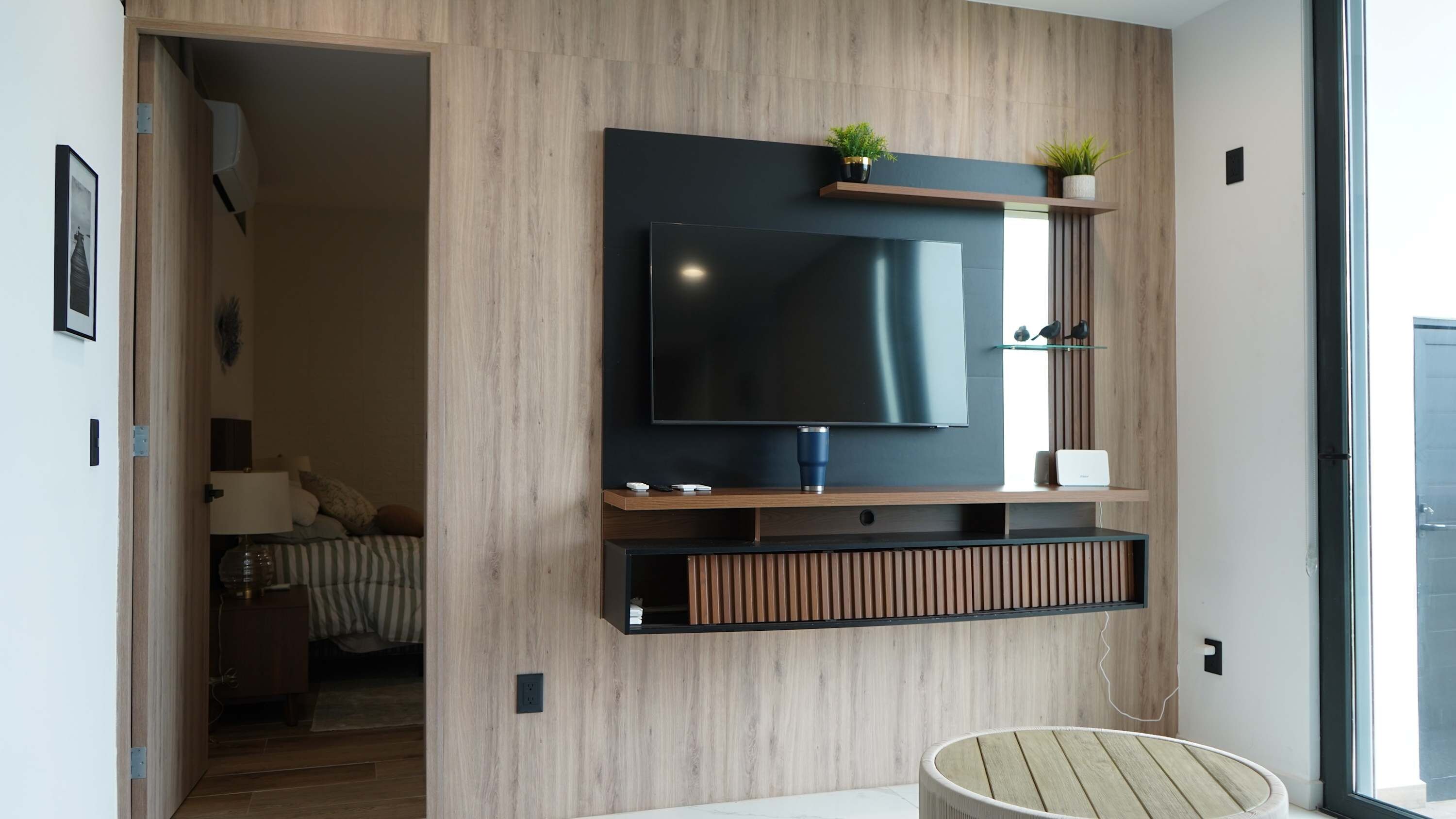 Un salon moderne avec un téléviseur à écran plat fixé à un mur en bois, une petite étagère ornée d’objets décoratifs et une fenêtre laissant entrer la lumière naturelle.