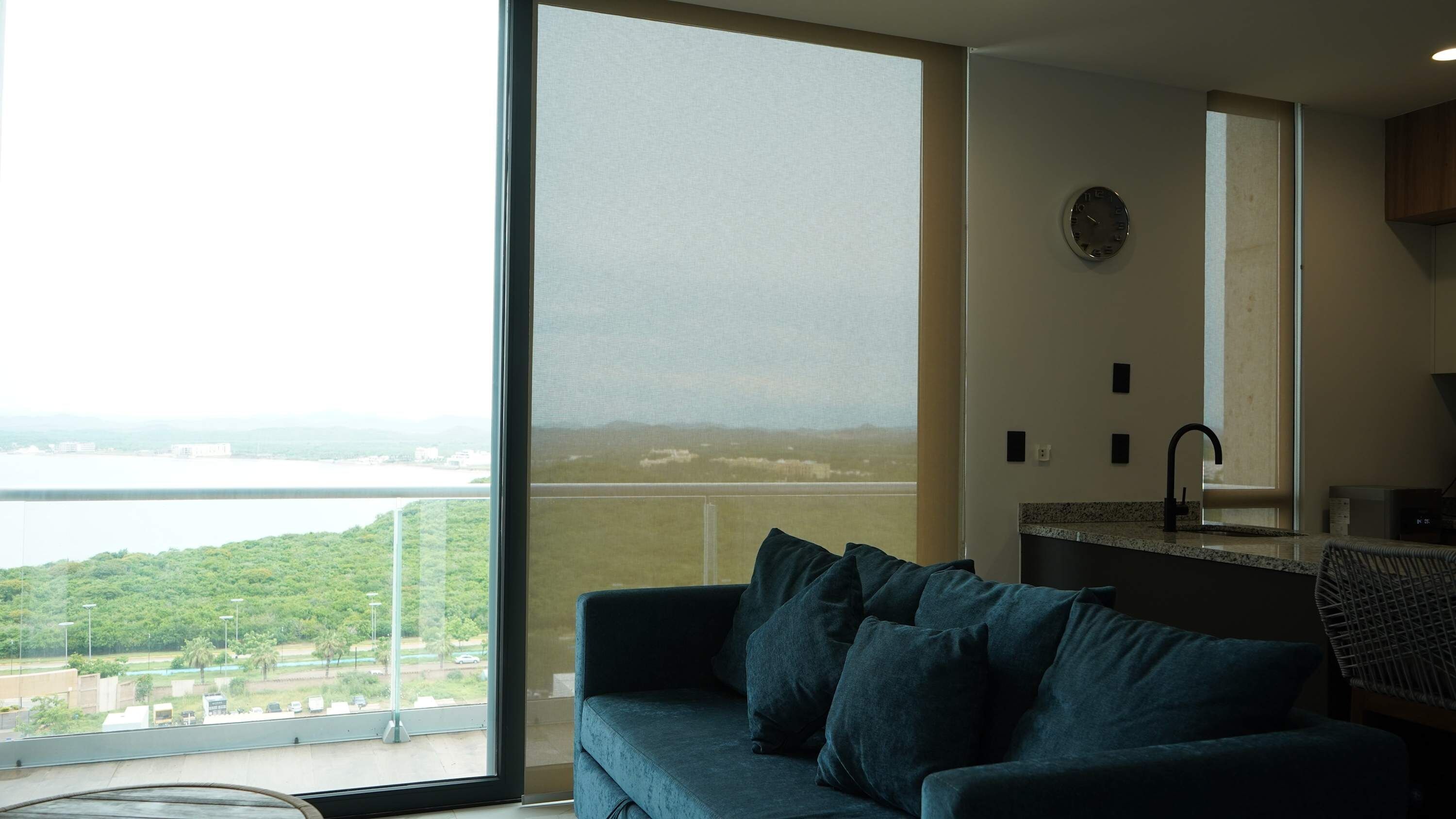 A room with a large window, a sofa, a kitchen area, and a view of a body of water and greenery.