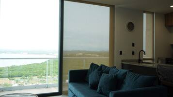 A room with a large window, a sofa, a kitchen area, and a view of a body of water and greenery.