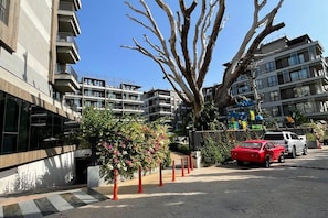 Eine moderne Stadtstraße mit mehrstöckigen Gebäuden, einem von Bäumen gesäumten Bürgersteig und einem roten Sportwagen, der am Straßenrand geparkt ist.