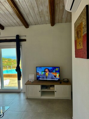 Ein Zimmer mit Fernseher auf einer Holzkommode, einem Glastisch und Blick auf ein Schwimmbad durch ein Fenster mit Vorhängen.