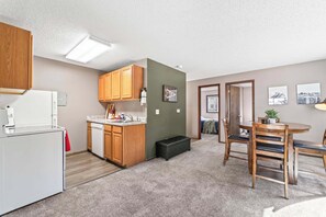 A compact kitchen with wooden cabinets, a dishwasher, and a refrigerator. There is a dining area with a table and chairs, and a hallway leading to another room. - 3BR 1BA, Sleeps 5 Walk to ISU (Ames)