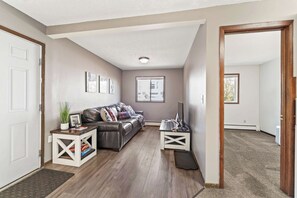 A living room with a leather sofa, a coffee table, and a TV stand. There are framed pictures on the wall and a door leading to another room. - 3BR 1BA, Sleeps 5 Walk to ISU (Ames)