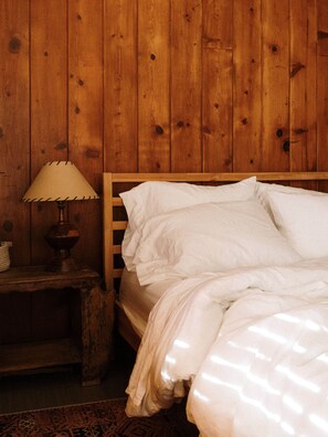 Ein hölzernes Bett mit weißer Bettwäsche in einem Zimmer mit Holzwänden und einem Nachttisch mit Lampe.