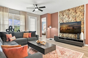 A modern living room with a sofa, coffee table, and a flat-screen TV mounted on a wall with a floral wallpaper. - Luxury 3BR, Newly Renovated, Lake View, Vista Cay (Orlando)