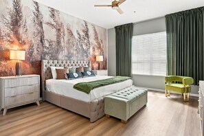 Une chambre à coucher avec un lit, des tables de chevet, un fauteuil vert, un ventilateur de plafond et une fenêtre avec des rideaux.