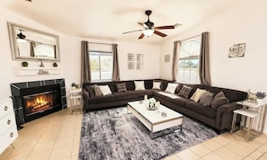 A modern living room with a sectional sofa, a fireplace, a ceiling fan, and a large window. - Serene Desert Oasis with Pool, Spa & Outdoor Kitchen (Desert Hot Springs)