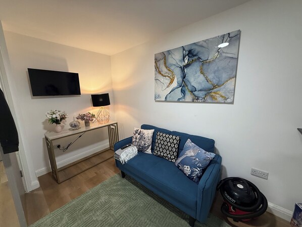 Ein Wohnzimmer mit einem blauen Sofa, einem Gemälde, einem Fernseher, einer Lampe und einem Staubsauger.
