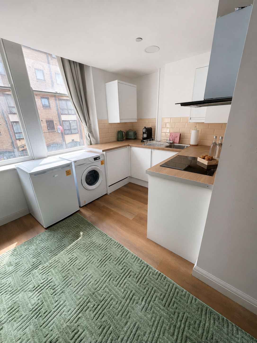 Une cuisine compacte équipée d’un lave-linge, d’un évier et d’une fenêtre donnant sur un immeuble.