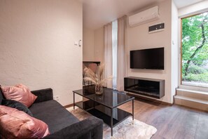 A modern living room with a flat-screen TV mounted on the wall, a glass coffee table, a dark sofa with pink cushions, and a large window with a view of greenery. - Kazinczy 11 - Boutique Apartment by BQA  (Budapest)