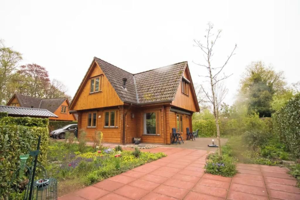 A wooden house with a sloped roof, a paved driveway, and a garden with various plants and flowers.