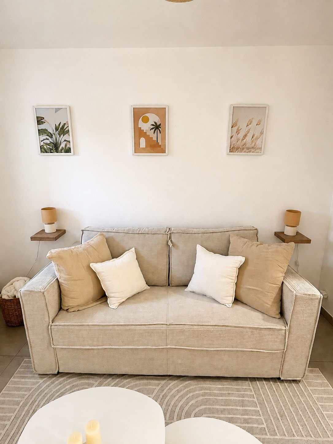 A neutral-toned living room with a beige sofa, two white cushions, and a round coffee table. There are three framed artworks on the wall above the sofa.