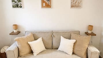 A neutral-toned living room with a beige sofa, two white cushions, and a round coffee table. There are three framed artworks on the wall above the sofa.