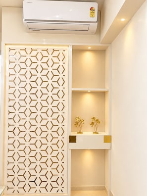 Una habitación con un equipo de aire acondicionado blanco, un separador decorativo con diseño geométrico y estantes con dos adornos dorados.