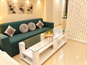 Una sala moderna con un sofá verde, cojines decorativos, una mesa de centro de vidrio con una planta y una pared con diseño geométrico.