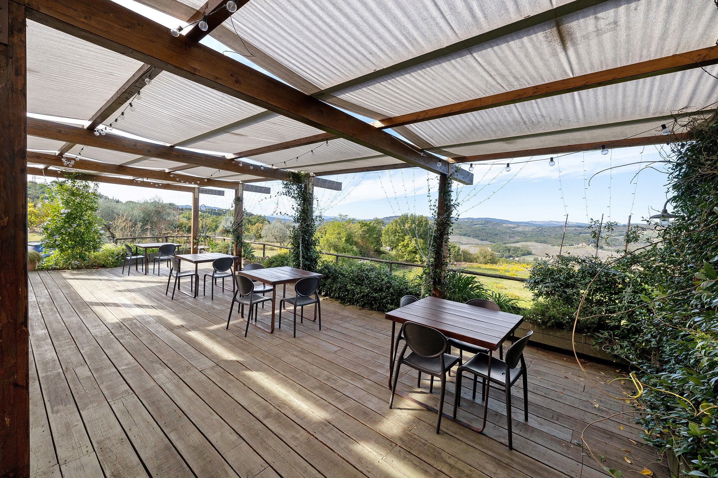 Un patio in legno con tavoli e sedie, un pergolato con tetto in metallo e vista sulla campagna.