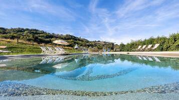 Ein klares Schwimmbecken mit Liegestühlen und Sonnenschirmen, umgeben von Grünflächen und einer hügeligen Landschaft.