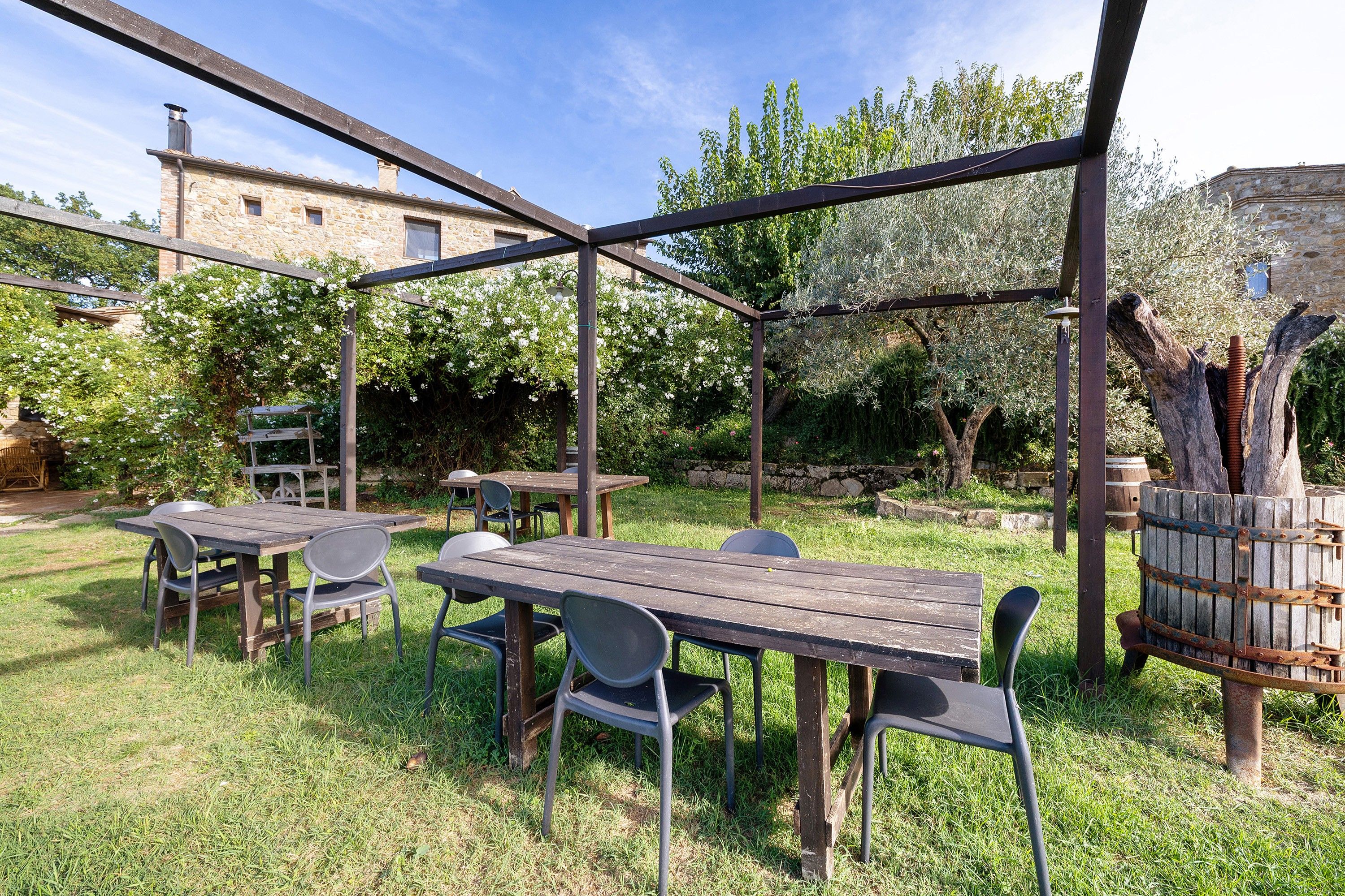 Un patio al aire libre con mesas y sillas de madera, una estructura de pérgola y un edificio de piedra al fondo.