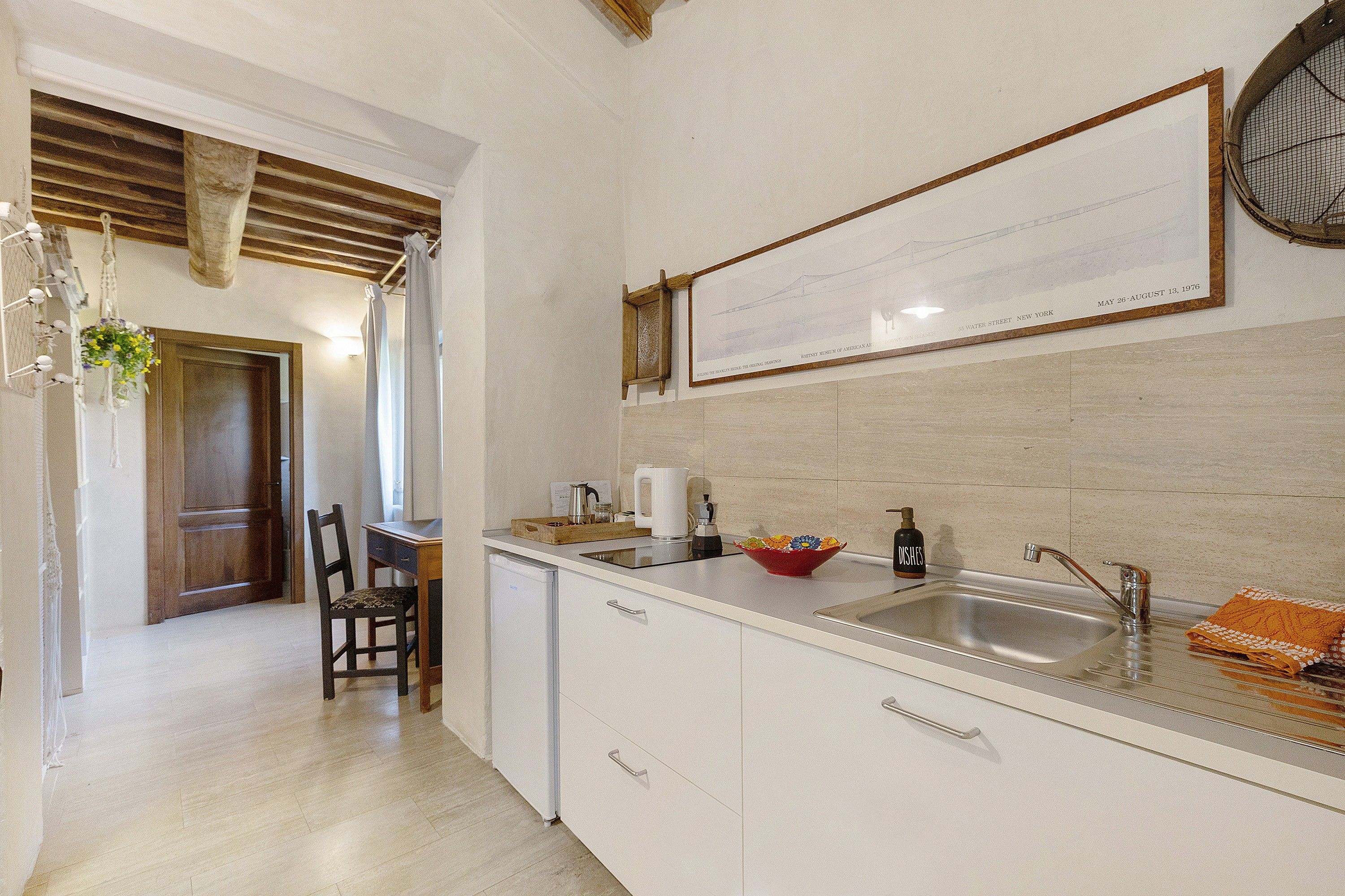 Una cocina moderna con lavavajillas integrado, un lavabo y una cafetera. Hay un cuadro en la pared y una ventana con cortinas.