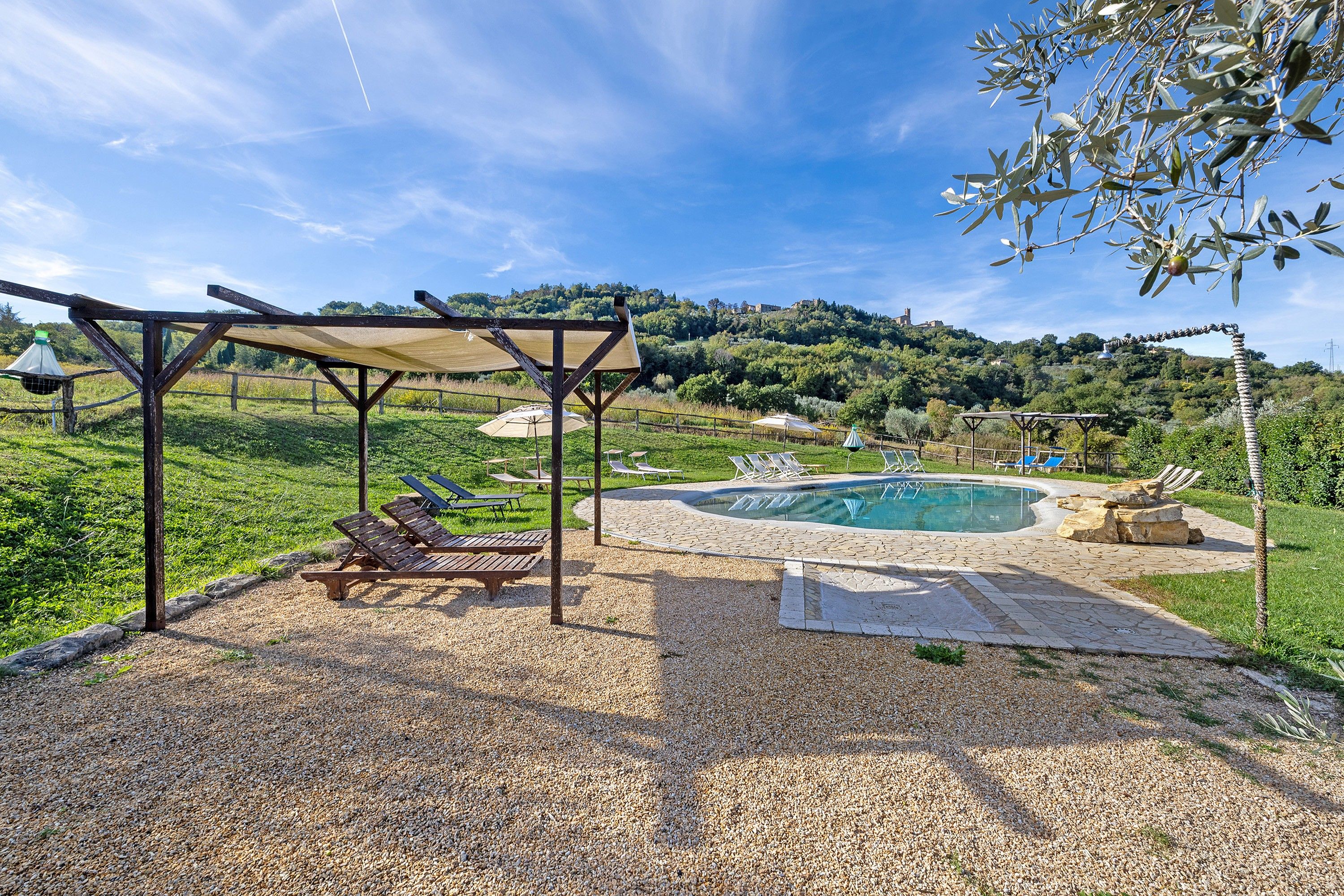 Area piscina con sedie a sdraio e un pergolato, circondata da un patio in ghiaia e vista su una collina.
