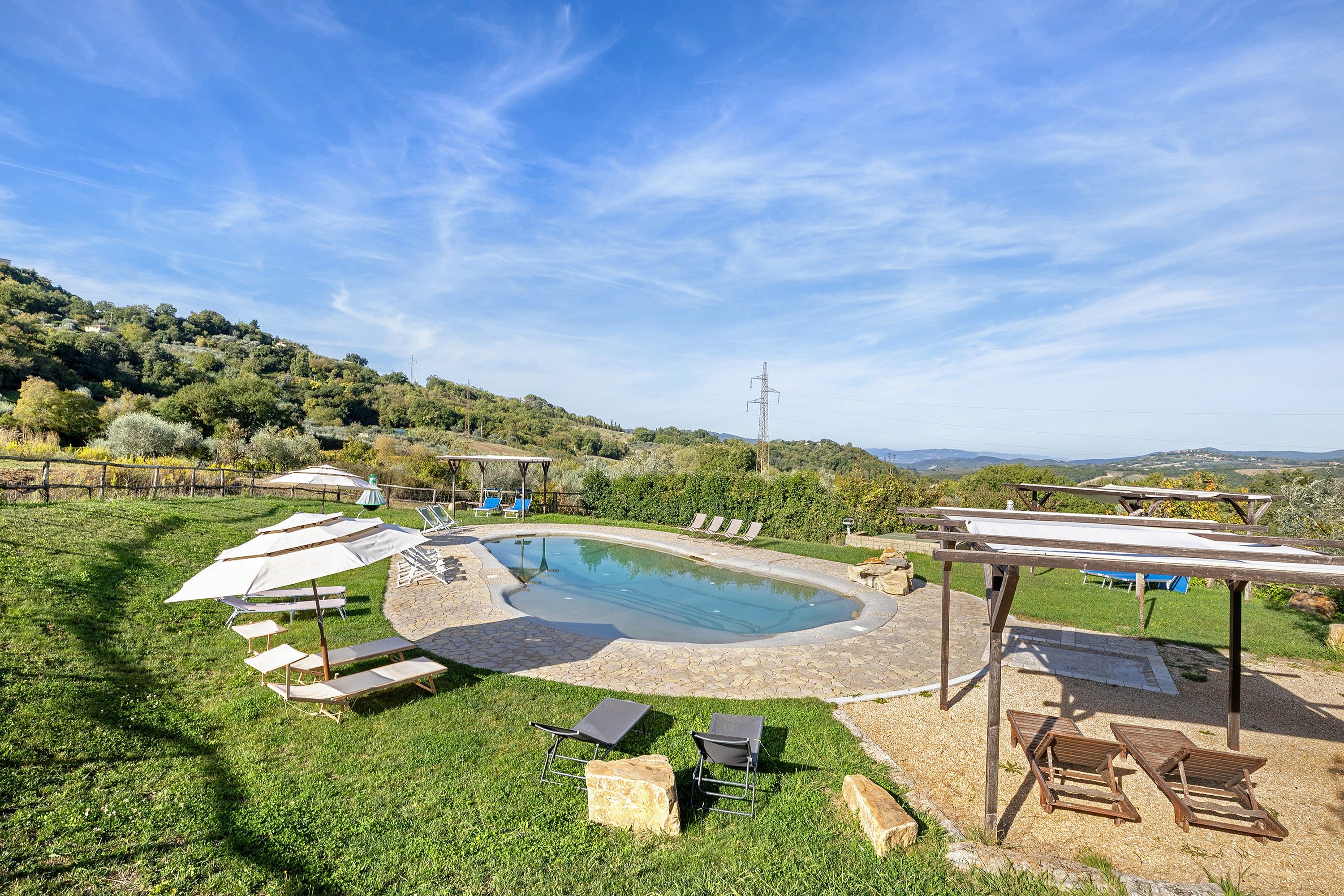 Una piscina con sillones de playa y sombrillas, rodeada de césped y colinas al fondo.