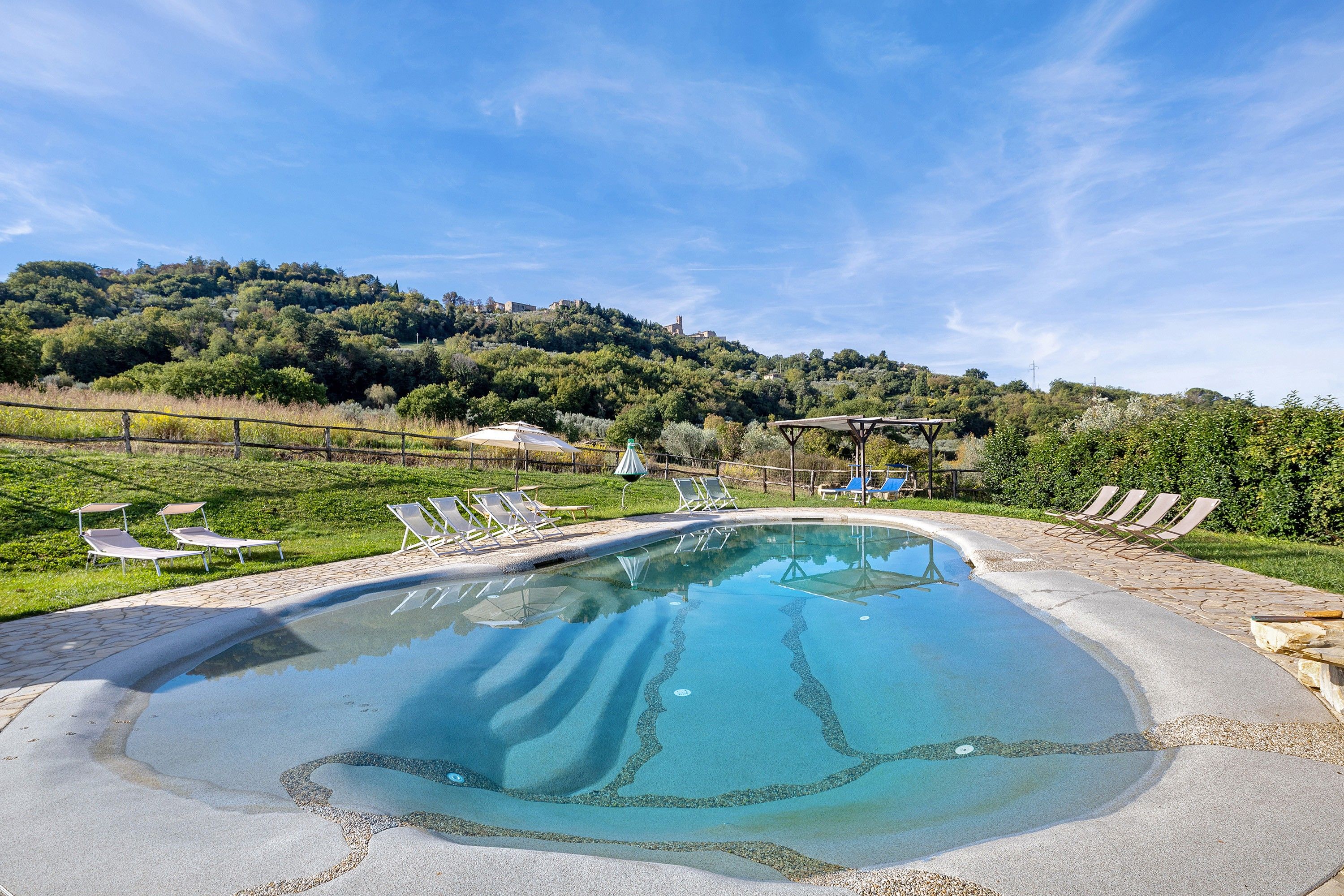Una piscina con sillones de descanso y una pérgola, rodeada de césped y colinas al fondo.