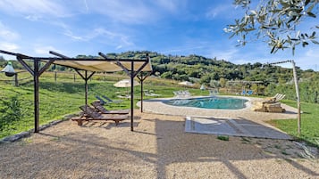Area piscina con sedie a sdraio e un pergolato, circondata da un patio in ghiaia e vista su una collina.