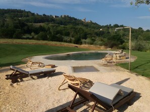 Ein Pool mit Liegestühlen und einer Whirlpool-Badewanne, umgeben von einem angelegten Garten und Blick auf einen Hügel mit Gebäuden.