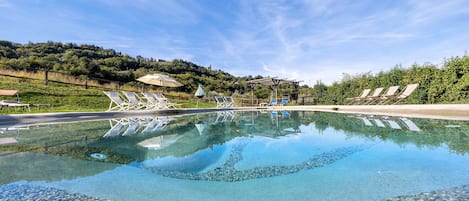 A clear swimming pool with lounge chairs and umbrellas, surrounded by greenery and a hilly landscape.