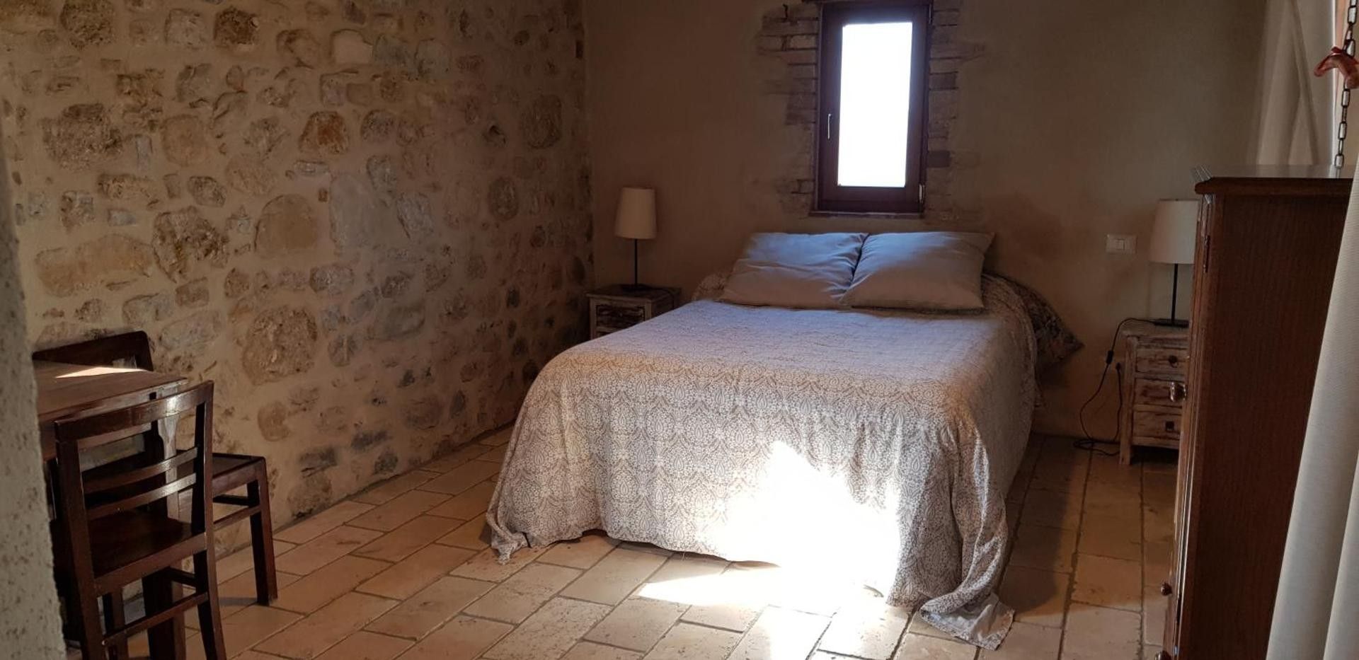 Ein Schlafzimmer mit Steinwand, einem Bett mit gemustertem Überwurf, einem Holzstuhl, einem Nachttisch mit Lampe und einem Fenster mit Tageslicht.