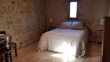 Ein Schlafzimmer mit Steinwand, einem Bett mit gemustertem Überwurf, einem Holzstuhl, einem Nachttisch mit Lampe und einem Fenster mit Tageslicht.