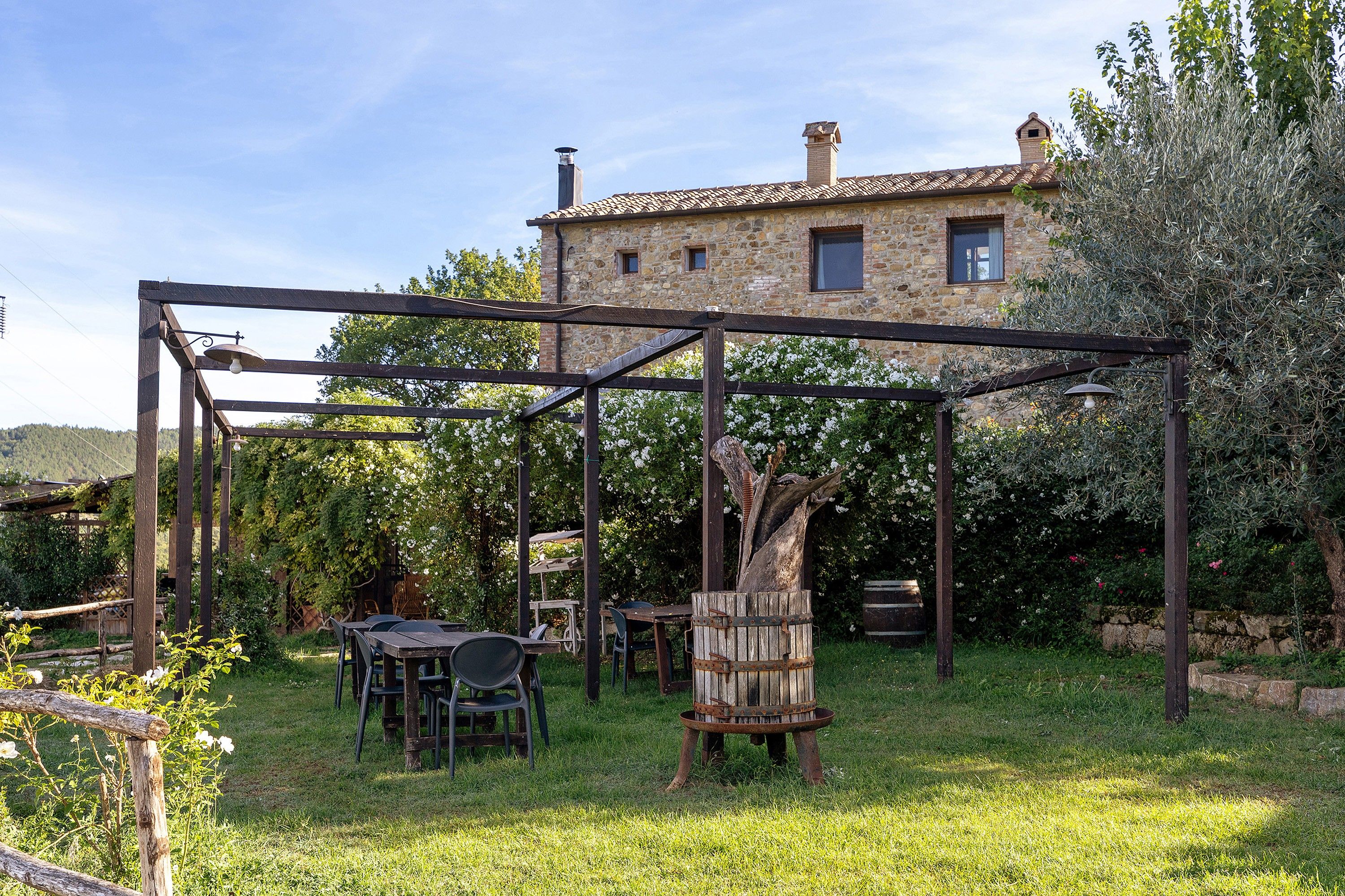 A rustic outdoor seating area with a pergola structure, stone building in the background, and a grassy lawn.