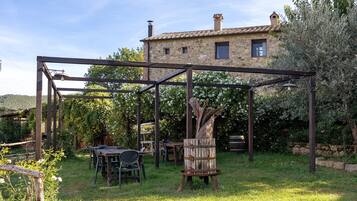 A rustic outdoor seating area with a pergola structure, stone building in the background, and a grassy lawn.