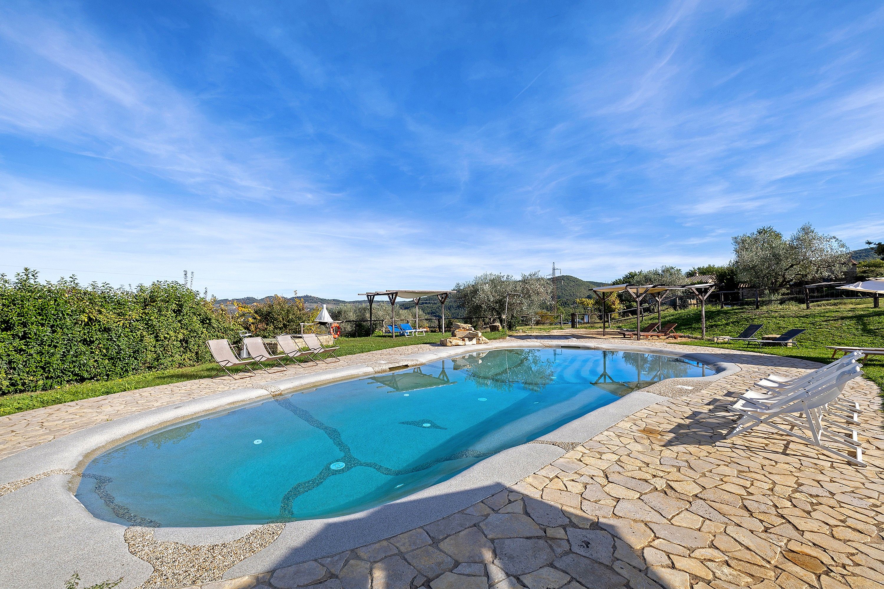 Una piscina con solárium de piedra, rodeada de vegetación y un cielo despejado.
