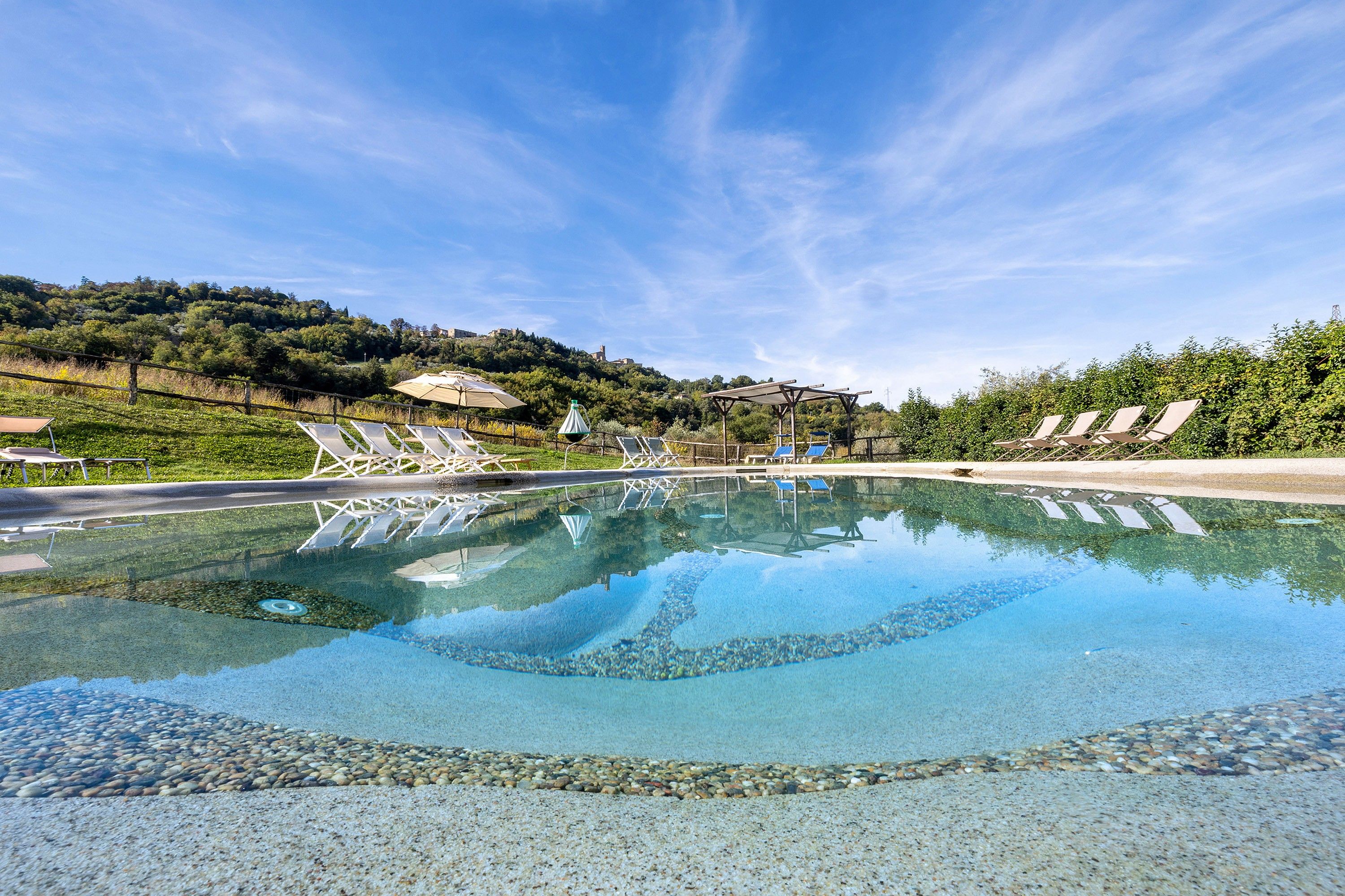Ein klares Schwimmbecken mit Liegestühlen und Sonnenschirmen, umgeben von Grünflächen und einer hügeligen Landschaft.