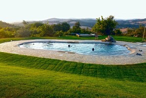 A circular hot tub with a stone border, surrounded by a well-maintained lawn and trees, set against a backdrop of hills. - Apartment 'Soffitte - Agriturismo Le Canterie' with Shared Pool, Private Terrace and Wi-Fi (Radicondoli)