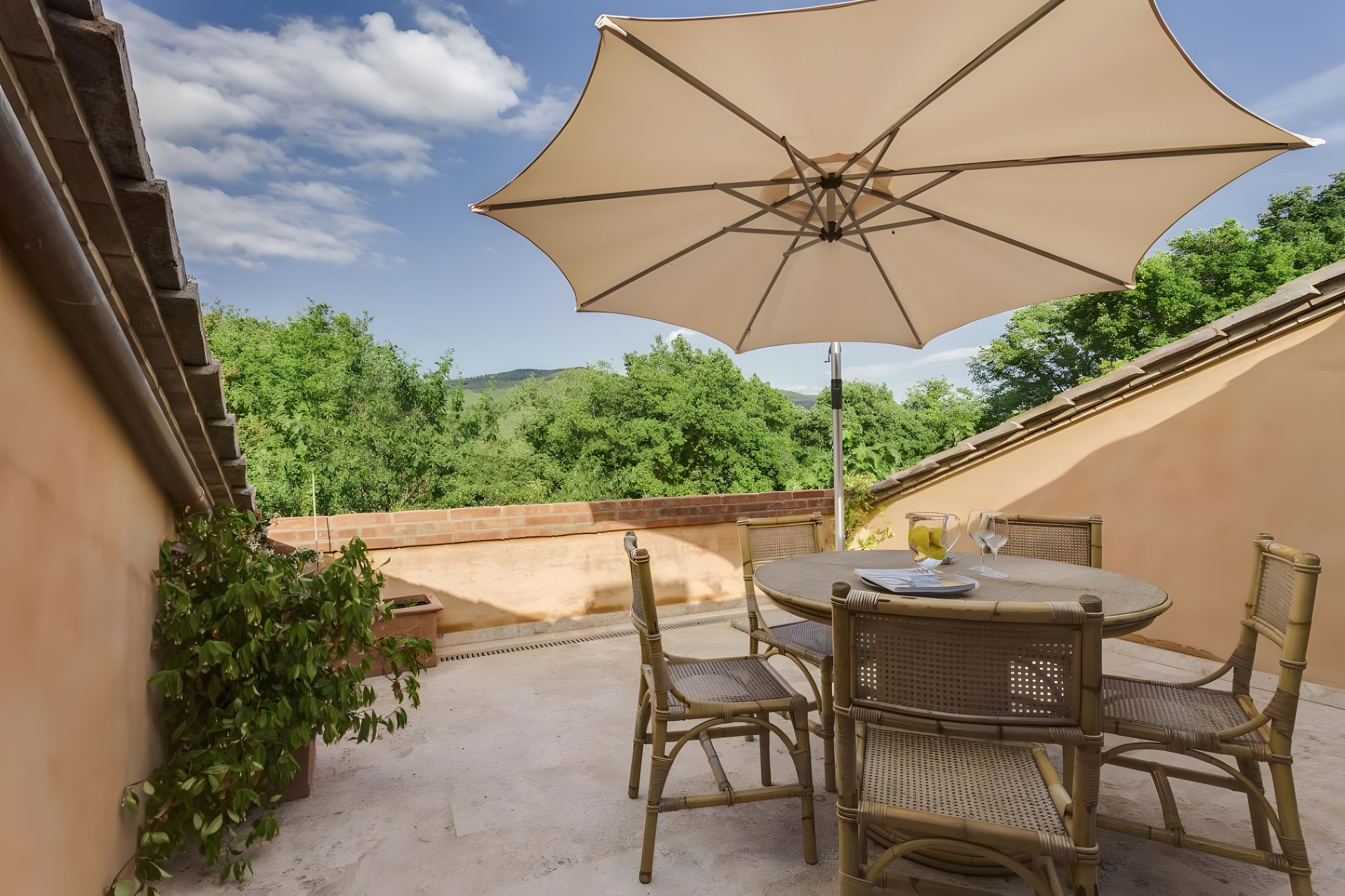 Eine Terrasse im Freien mit Tisch und Stühlen, einem großen Sonnenschirm und Blick auf Grünflächen und Hügel.