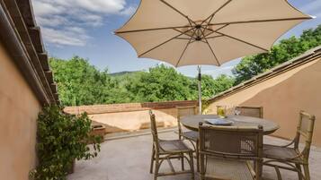 Eine Terrasse im Freien mit Tisch und Stühlen, einem großen Sonnenschirm und Blick auf Grünflächen und Hügel.