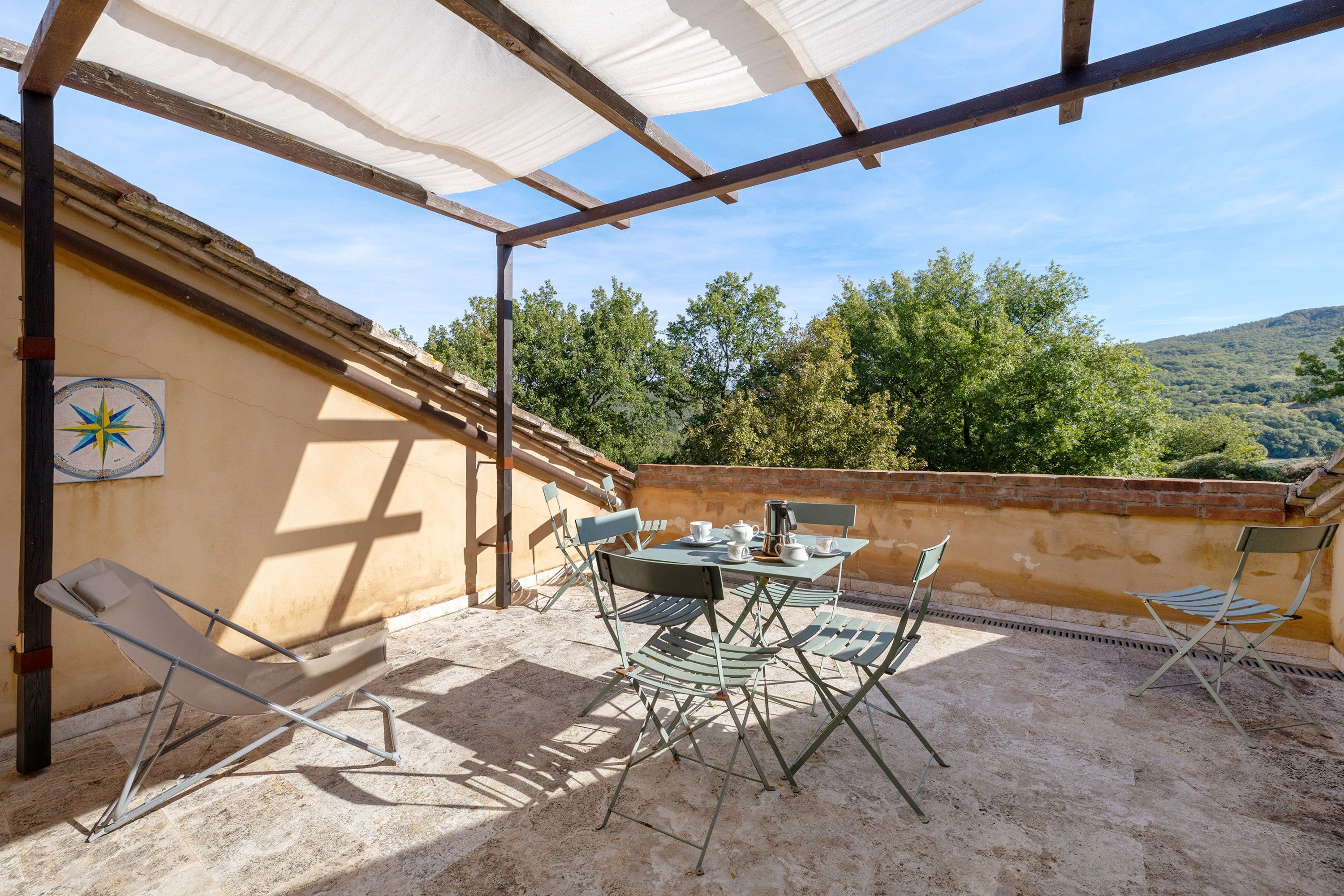 Eine überdachte Terrasse im Freien mit Tisch und Stühlen, Sonnenschirm und Blick auf einen bewaldeten Hügel.