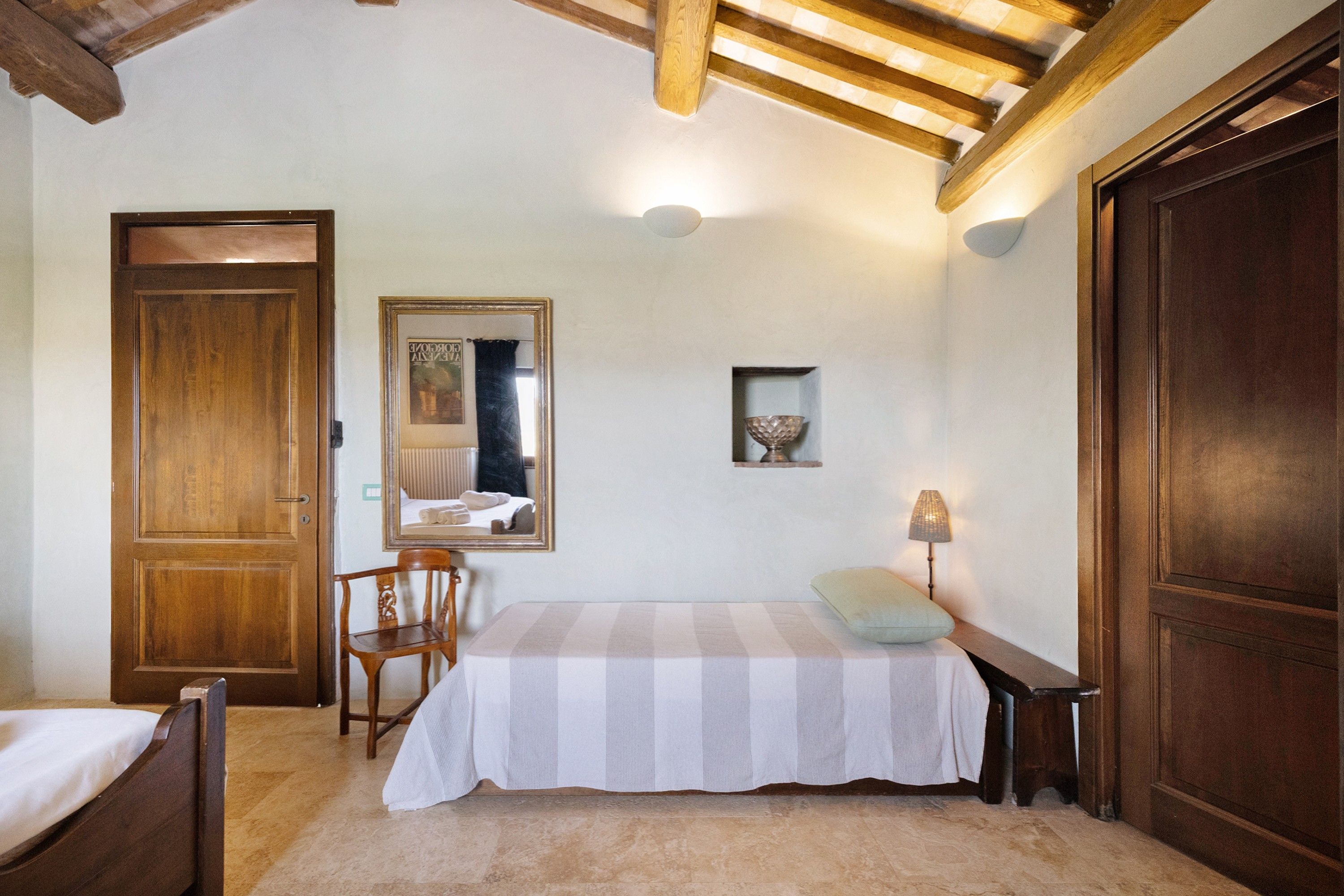 Un dormitorio con cama, una silla, puerta de madera, espejo, mesita de noche con lámpara y una pequeña estantería con un cuenco.