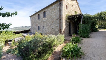 Un edificio de piedra con techo inclinado, camino de grava y jardín con diversas plantas.