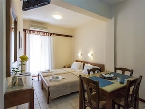Una habitación de hotel con una cama, una mesa de comedor, sillas, un florero con flores y una ventana con cortinas.