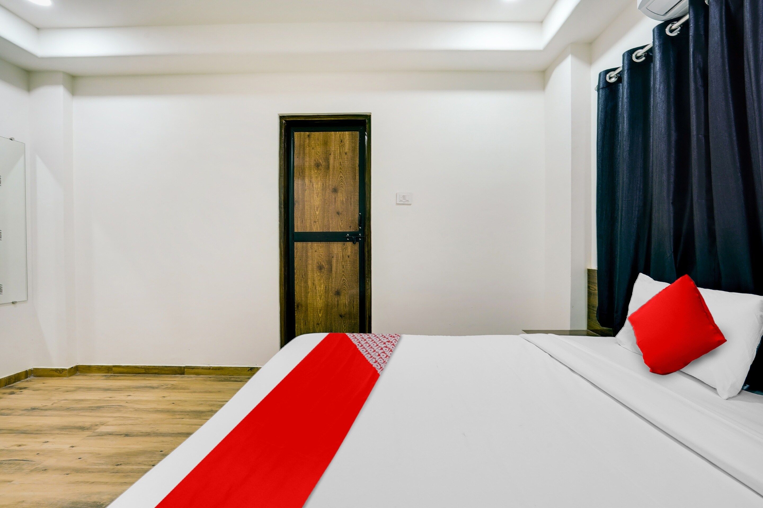 A hotel room with a wooden door, a bed with white bedding and a red pillow, and a dark curtain.