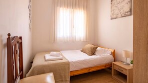 Ein Einzelbett mit Holzgestell, ein Nachttisch mit Lampe und ein Fenster mit leichtem Vorhang.