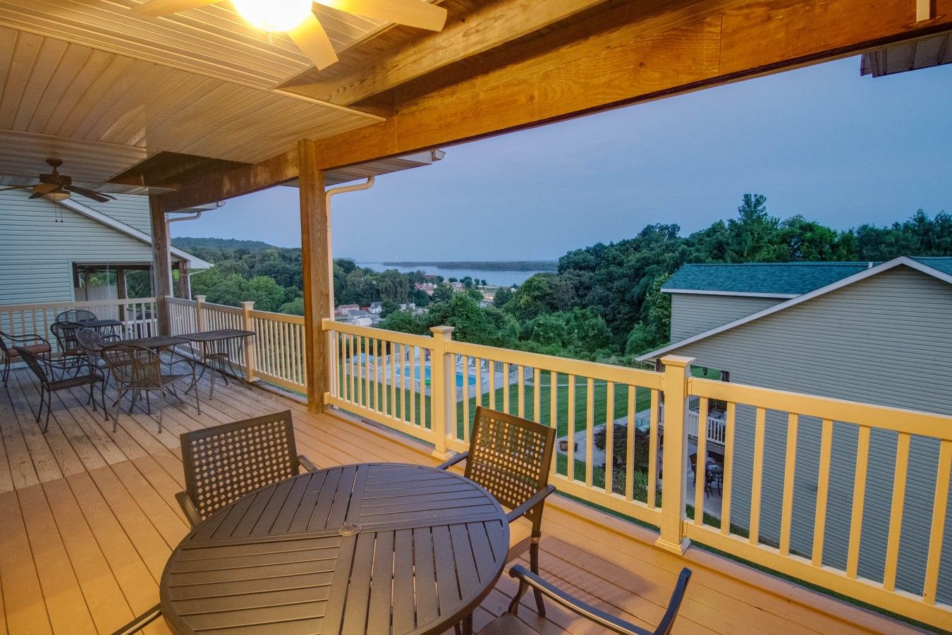 A covered outdoor deck with a table and chairs, a view of a body of water, and a green landscape.