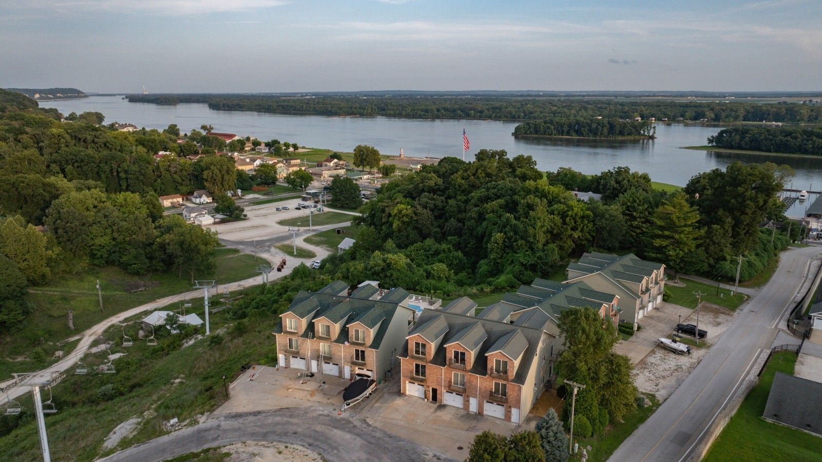 A residential area with a large building featuring a brick facade and multiple garage doors, surrounded by greenery and a body of water.