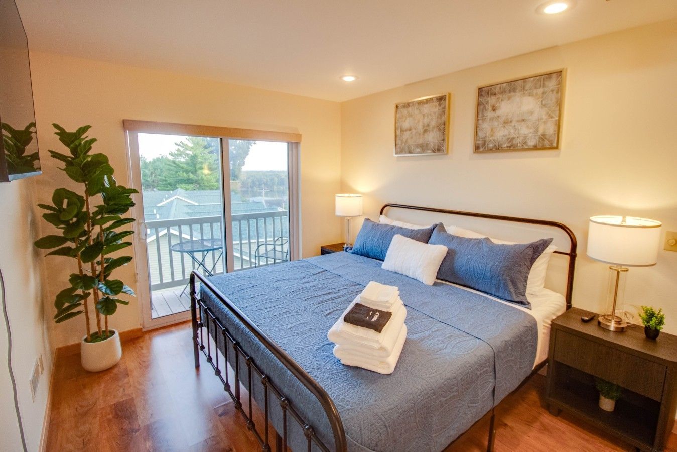 A hotel room with a bed, a balcony, a potted plant, and artwork on the wall.